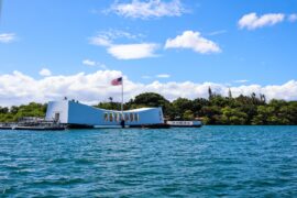 The USS Arizona Memorial is over the water in Pearl Harbor. An American flag on a flagpole waves over the building.