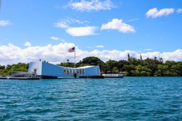 The USS Arizona Memorial is over the water in Pearl Harbor. An American flag on a flagpole waves over the building.