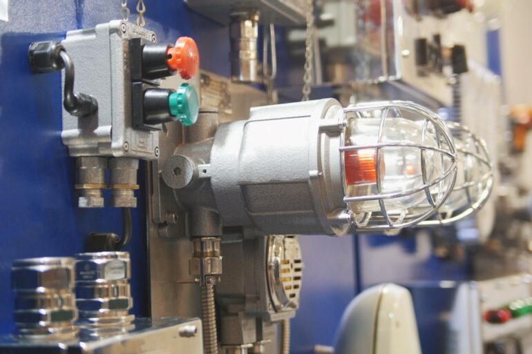 An industrial gas leak control panel and fire alarm light with a protective cage mounted on equipment in a manufacturing facility.