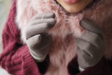 A person wearing a knitted wine-colored sweater, a furry pink scarf, and muted brown suede-like winter gloves.