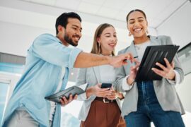 Three coworkers smile over a tablet in a bright office; one points while others hold a phone and notebook.