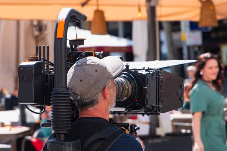 A camera operator films an actress at an outdoor cafe during a remote film shoot using a professional camera rig.