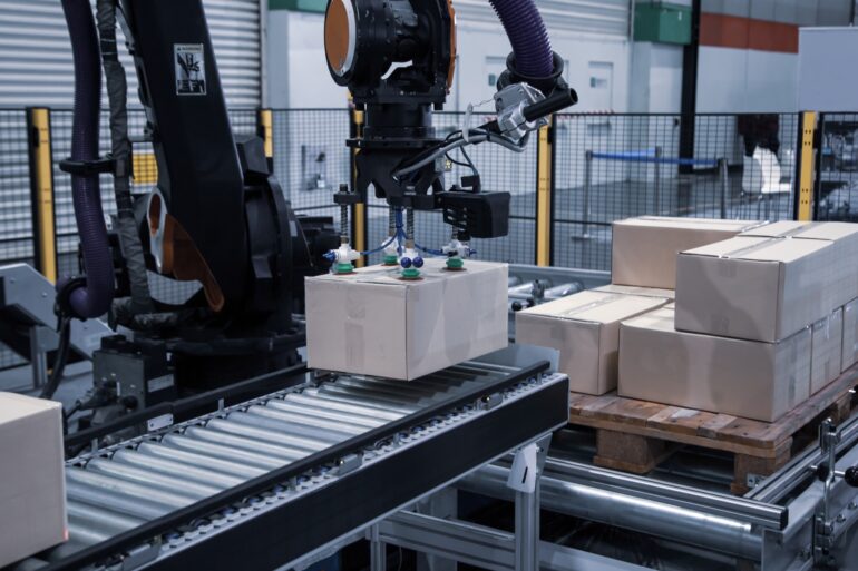 A robotic arm with a suction gripper lifts a cardboard box from a conveyor belt onto a pallet in an automated warehouse line.
