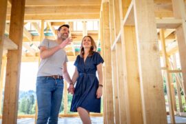 A man and a woman holding hands while walking inside a hallway of a home under construction with wooden beams.