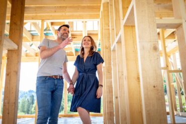 A man and a woman holding hands while walking inside a hallway of a home under construction with wooden beams.