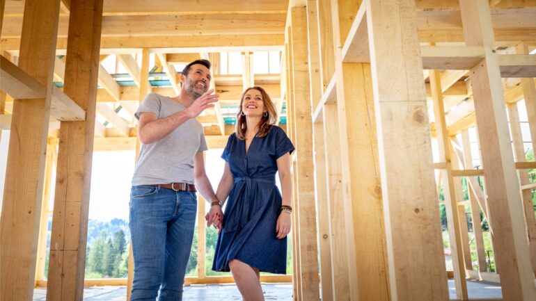 A man and a woman holding hands while walking inside a hallway of a home under construction with wooden beams.