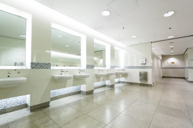 A clean and modern public restroom features seven white sinks attached to a tan-tiled wall. Four mirrors are on the wall.