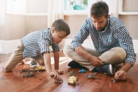 A man and a young boy in matching khakis and checkered shirts sit on the floor, playing with toy cars and action figures.