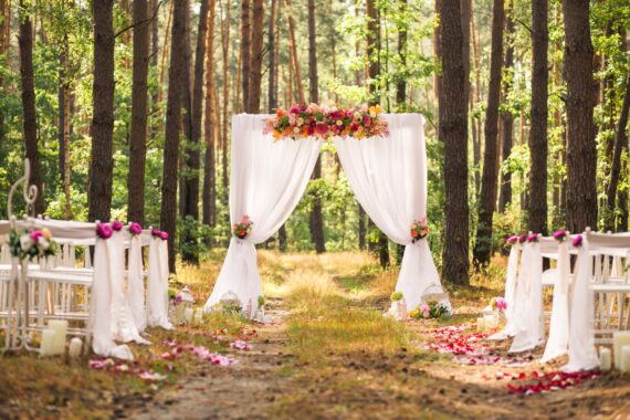 A decorated outdoor wedding setup in a forest features a white arch with flowers, chairs, and a petal-lined aisle.