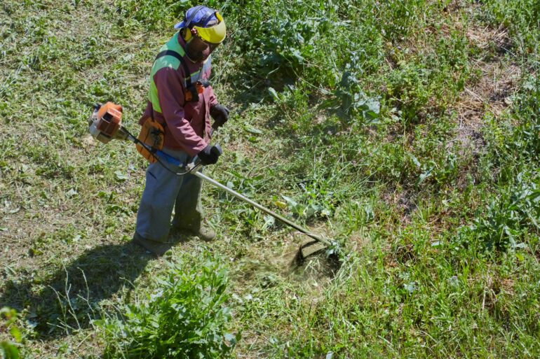 An aerial view of a man in full protective gear, using a weedwhacker to clear an overgrown section of yard.