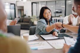 Five business professionals sitting around a table with papers. The woman is shaking hands with the man across from her.