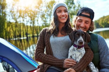 A man and a woman are sitting outside with their puppy. A body of water and a bank of trees are in the background.