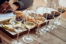 A flight of six wine tastings, three white and three red, lined up next to a charcuterie board with bread, cheese, fruit, and dip.
