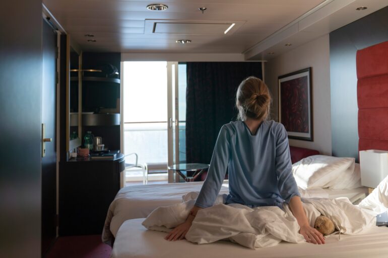 Person sits on unmade bed in modern hotel room, facing bright balcony doors with morning light entering space.