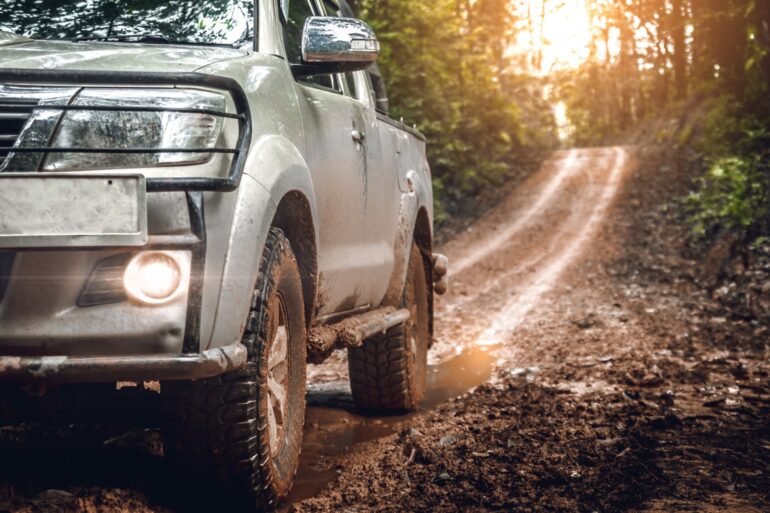 The driver's side of a silver truck driving along a muddy trail. Mud coats the tires, and the sun is behind the trees.