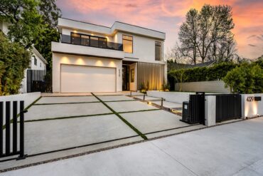 A two-story house has its outside lights on at dusk. It features a very large concrete driveway and a metal gate.