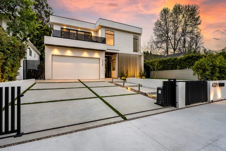 A two-story house has its outside lights on at dusk. It features a very large concrete driveway and a metal gate.
