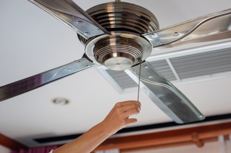 A hand reaches over to pull the cord on a metal ceiling fan in a room. The device hangs from a white ceiling.