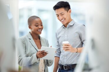 A businesswoman holds a tablet and shows the screen to a coworker who's holding a coffee. They're standing in an office.