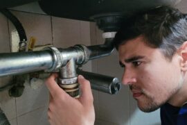 A man kneeling under a sink while inspecting a pipe connection with hands positioned near the plumbing components.