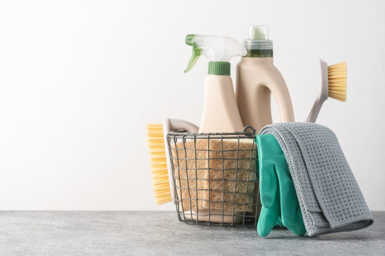 A wire basket holds eco-friendly cleaning supplies including refillable bottles, brushes, sponges, and a cleaning cloth.