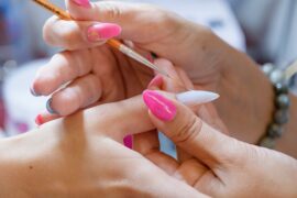 A close-up of a manicurist applying nail art to a client’s fingernails using a small brush while holding the hand steady.