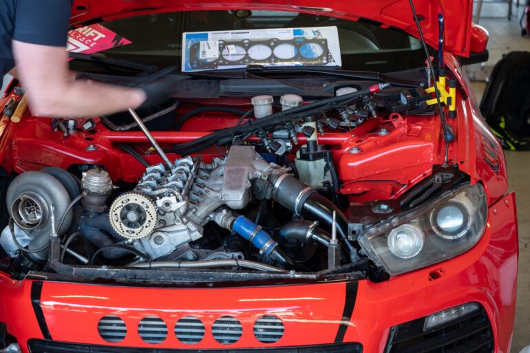 A person stands beside an open car hood, showcasing its large and intricately modified engine in a garage.