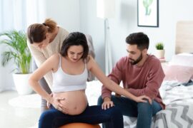 A pregnant woman sits on an orange yoga ball as a woman stands behind her, massaging her back. A man holds her hand.