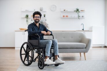 A wheelchair user in a well-lit living room with a sofa behind him. There are also shelves on the wall and a counter.