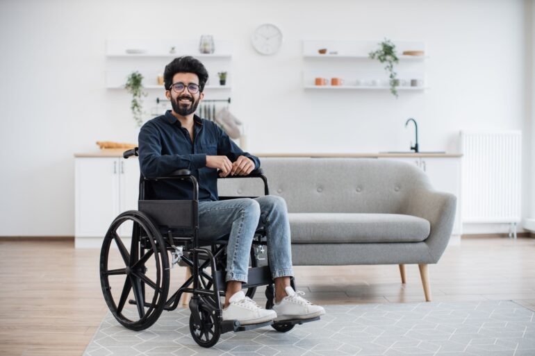 A wheelchair user in a well-lit living room with a sofa behind him. There are also shelves on the wall and a counter.