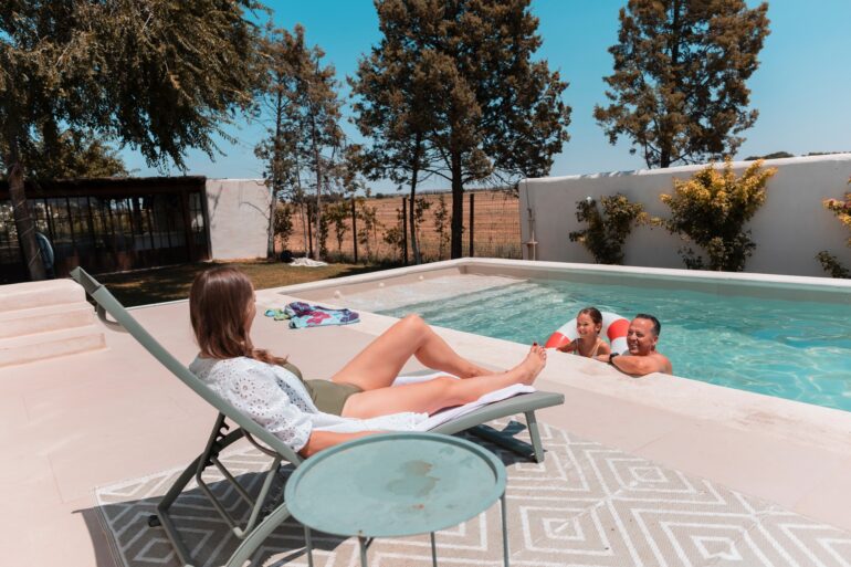 Family members enjoying nice weather in a home pool. One of them is lying in a beach chair in the sun.