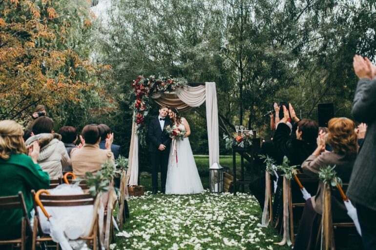 A bride and groom stand beneath a floral arch during an outdoor wedding ceremony as seated guests applaud.
