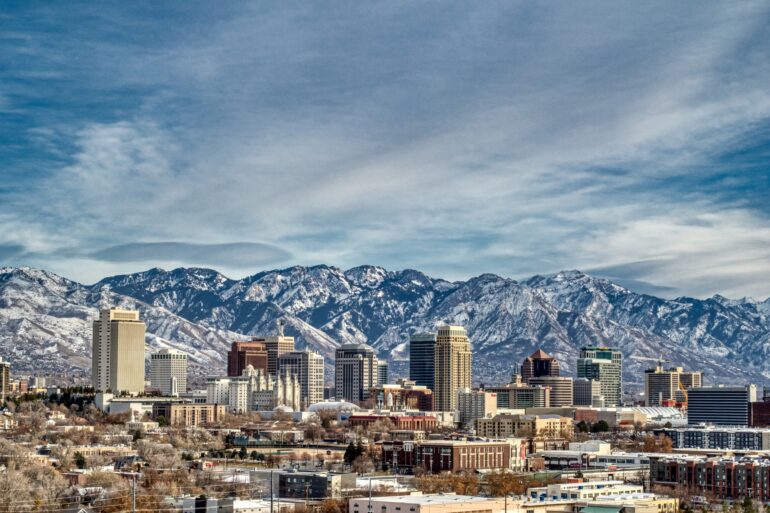 A landscape view of the skyline of Salt Lake City, including skyscrapers and snow-capped mountains behind them.
