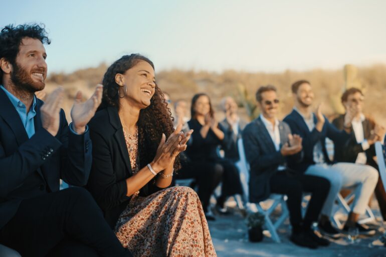 A bunch of people seated outdoors in rows of chairs for a wedding. They are all currently smiling while clapping.