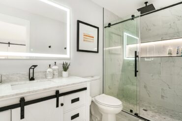 A modern bathroom with white cabinets, black hardware, a walk-in shower with large glass doors next to a toilet.