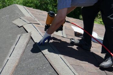 A close-up of the hands of a man wearing safety gloves, using a nail gun to attach a shingle on the top of a roof.