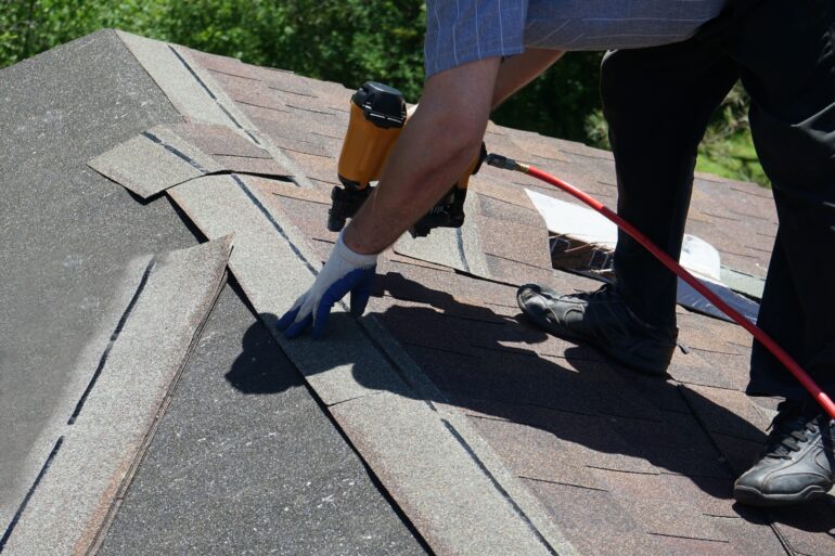 A close-up of the hands of a man wearing safety gloves, using a nail gun to attach a shingle on the top of a roof.