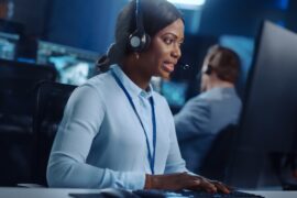 A woman wearing a blue blouse and a black headset, sitting behind a desk, typing on a keyboard next to a monitor.