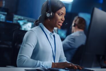 A woman wearing a blue blouse and a black headset, sitting behind a desk, typing on a keyboard next to a monitor.