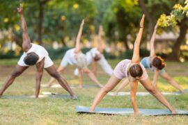 Five people are doing yoga on yoga mats in a grassy area outside. Trees appear blurry behind them.