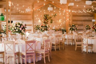 A rustic wedding reception with glowing string lights, round tables, and floral centerpieces in a wood venue.