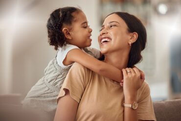 Smiling woman holds a young child with braided hair as they laugh together in a softly lit home setting.