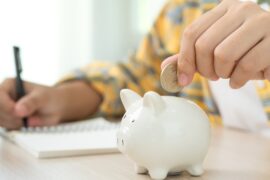 A close-up view shows a person placing a coin into the opening of a white piggy bank as they write in a notepad.
