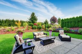 A patio with wicker chairs, a table, and potted plants sitting in front of a beautifully landscaped backyard.