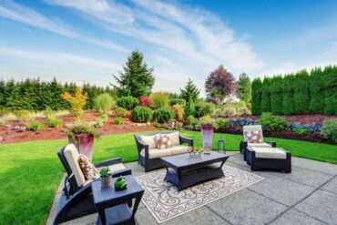 A patio with wicker chairs, a table, and potted plants sitting in front of a beautifully landscaped backyard.