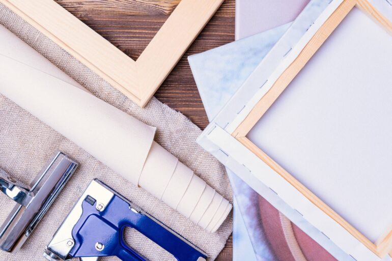 A blue staple gun, rolled fabric, and wooden canvas frames arranged beside folded canvas material on a brown wood surface.