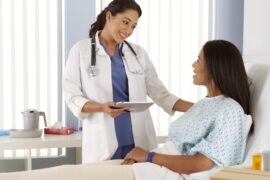 A woman lies in a hospital bed, wearing a gown. A doctor holding a tablet touches the patient's shoulder.