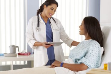 A woman lies in a hospital bed, wearing a gown. A doctor holding a tablet touches the patient's shoulder.
