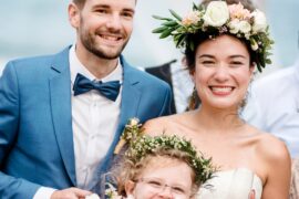 A smiling couple and child in wedding attire stand beside one another. The bride and child wear flower crowns.