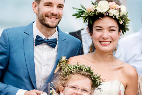 A smiling couple and child in wedding attire stand beside one another. The bride and child wear flower crowns.